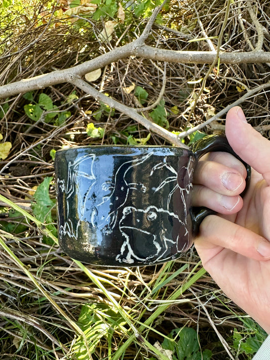 A hand holding a glossy black ceramic mug with white line-art illustrations of animals, set against a natural outdoor background of dry grass and branches.