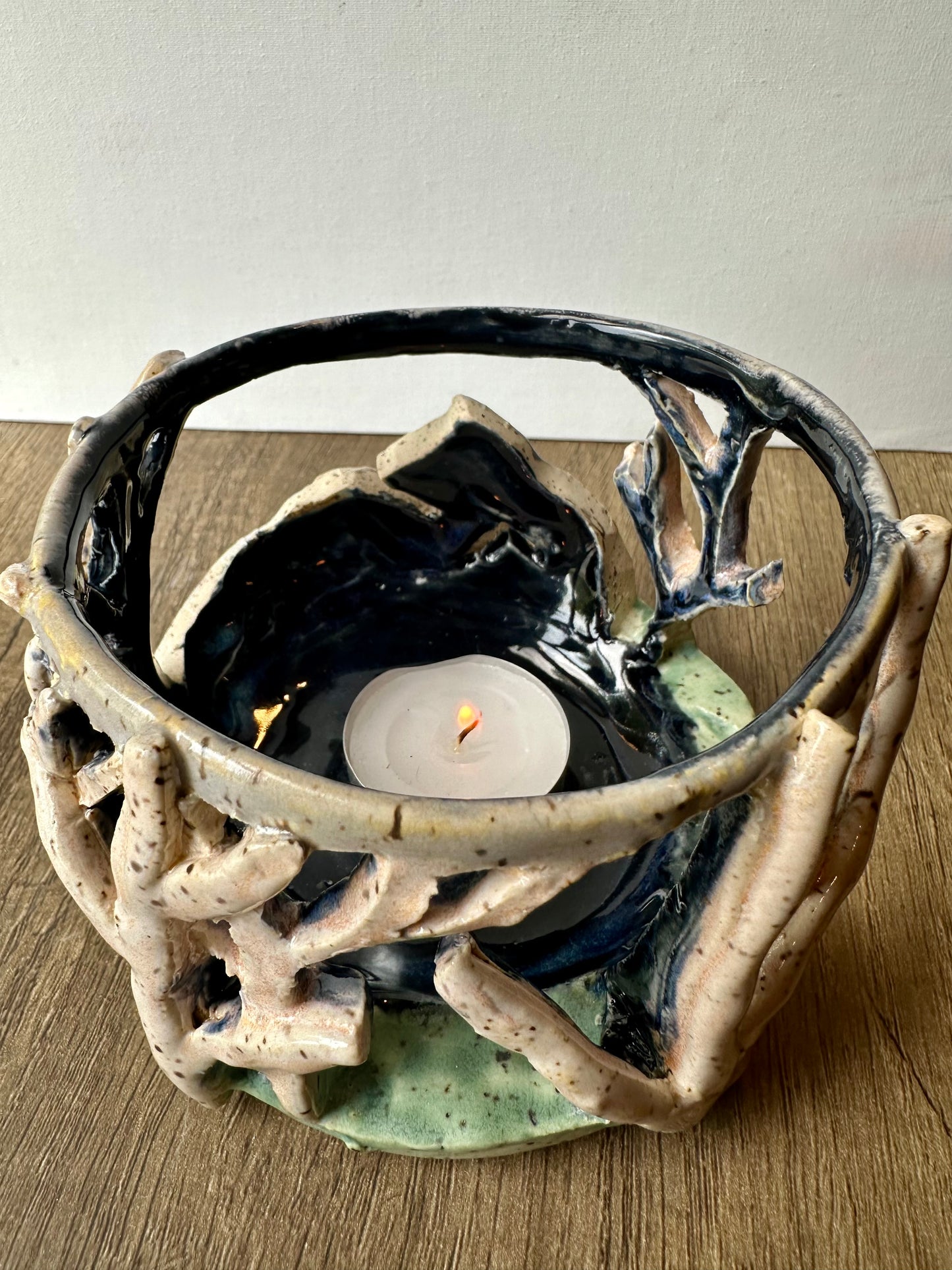 Round ceramic candle surround with mountain and birch trees cut out; lit tea light candle placed inside.