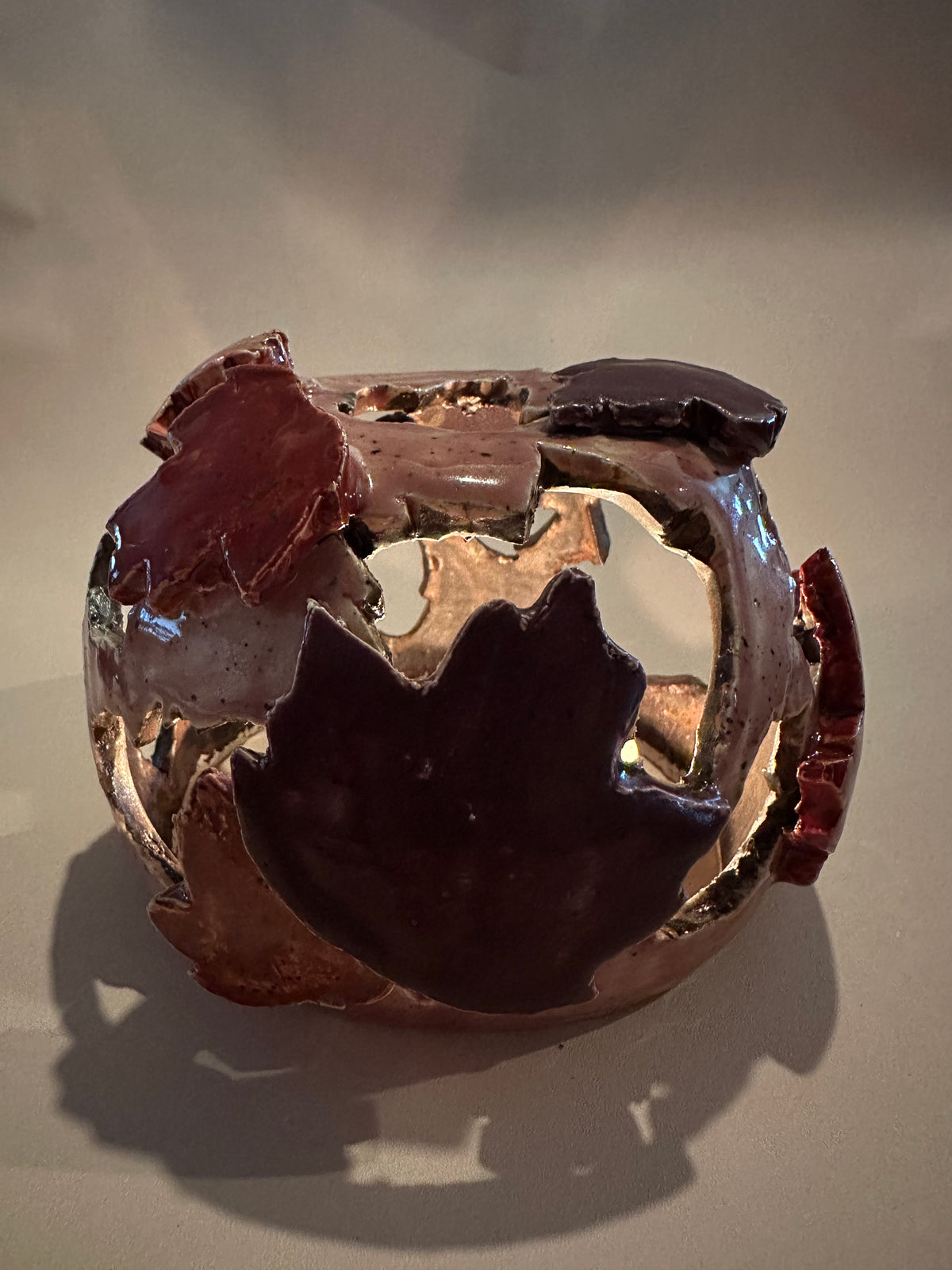 A decorative ceramic sphere with glossy mauve base glaze, featuring maple leaf-shaped cutouts in various shades of red, with three lit tea lights inside, on a neutral beige background.