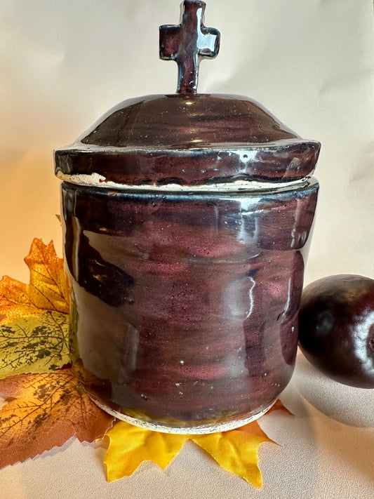 A glossy black canister topped with a lid with a cross as a handle. Styled with fall leaves and an apple on a solid neutral background.