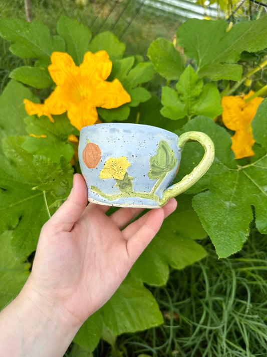 A hand holds a blue ceramic mug with illustrations of pumpkins, pumpkin flowers, pumpkin leaves, and a green handle; set against a garden background of pumpkin leaves and pumpkin flowers.