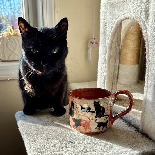 A black cat with green eyes sits on a cat tree next to a ceramic mug decorated with black and orange cat silhouettes, in a brightly lit room.
