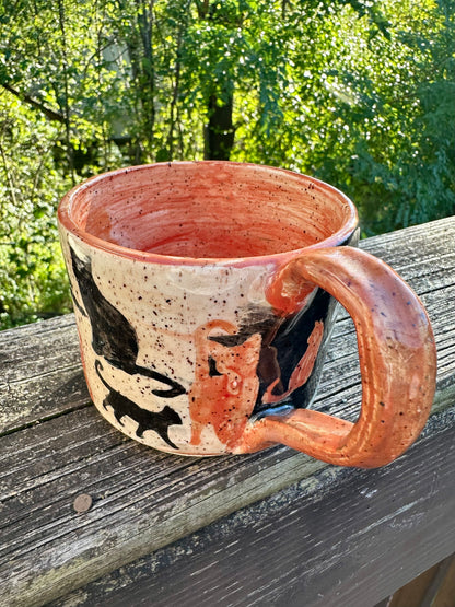 A handmade ceramic mug with an exterior transitioning from white to black, decorated with cat silhouettes in black and orange, featuring an orange handle and interior, set on a weathered wooden railing with a lush green forest background.