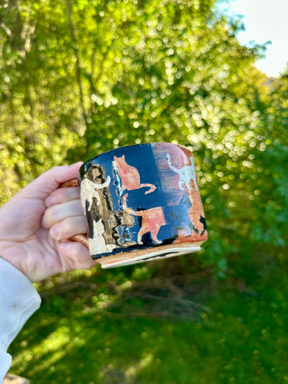 A hand holding a handmade ceramic mug with an exterior transitioning from black to orange with orange, white, and black painted cat illustrations; set against a blurred green outdoor background.