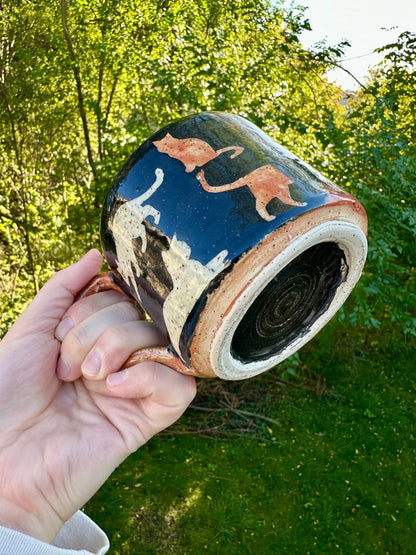 A hand holding a handmade ceramic mug with a black exterior with orange, white, and black painted cat illustrations and an orange, white, and black bottom; set against a blurred green outdoor background.