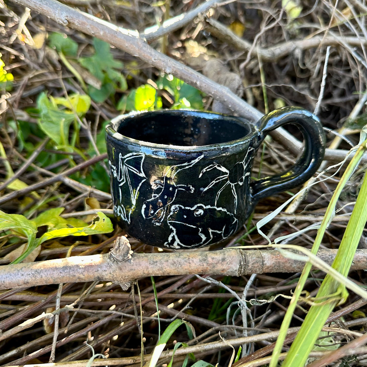 A small black ceramic cup with white line-art illustrations of figures and animals, placed outdoors on a branch surrounded by dry twigs and green leaves.