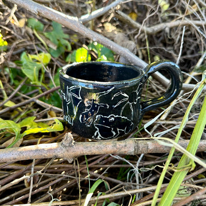 A small black ceramic cup with white line-art illustrations of figures and animals, placed outdoors on a branch surrounded by dry twigs and green leaves.