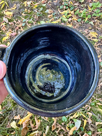 Interior shot of a handmade glossy black glazed ceramic mug with a subtle sparkle, held by a hand against a background of grass and fallen autumn leaves.
