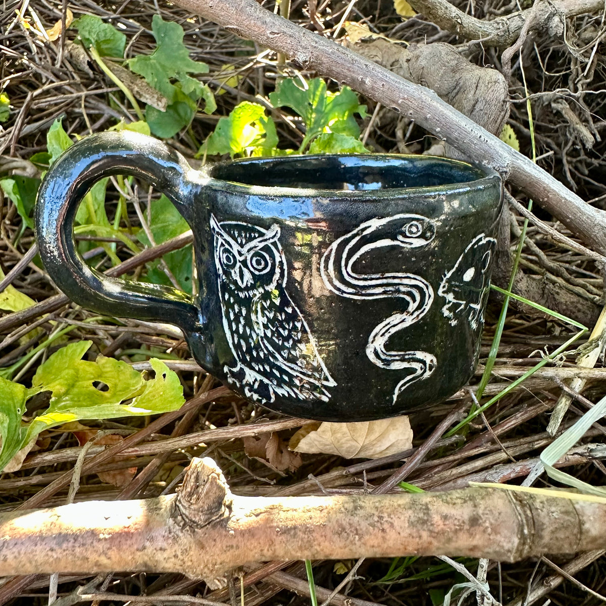 A small black ceramic cup with white line-art illustrations of figures and animals, placed outdoors on a branch surrounded by dry twigs and green leaves.