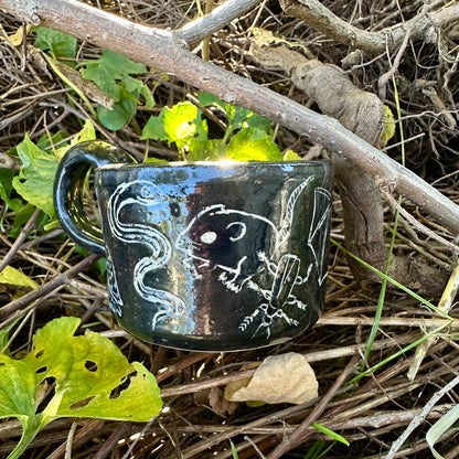 A small black ceramic cup with white line-art illustrations of figures and animals, placed outdoors on a branch surrounded by dry twigs and green leaves.