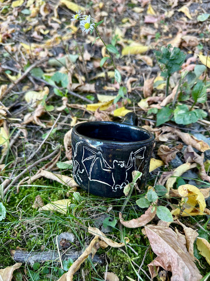 A small black ceramic cup with white line-art illustrations of figures and animals, placed outdoors on a branch surrounded by dry twigs and green leaves.