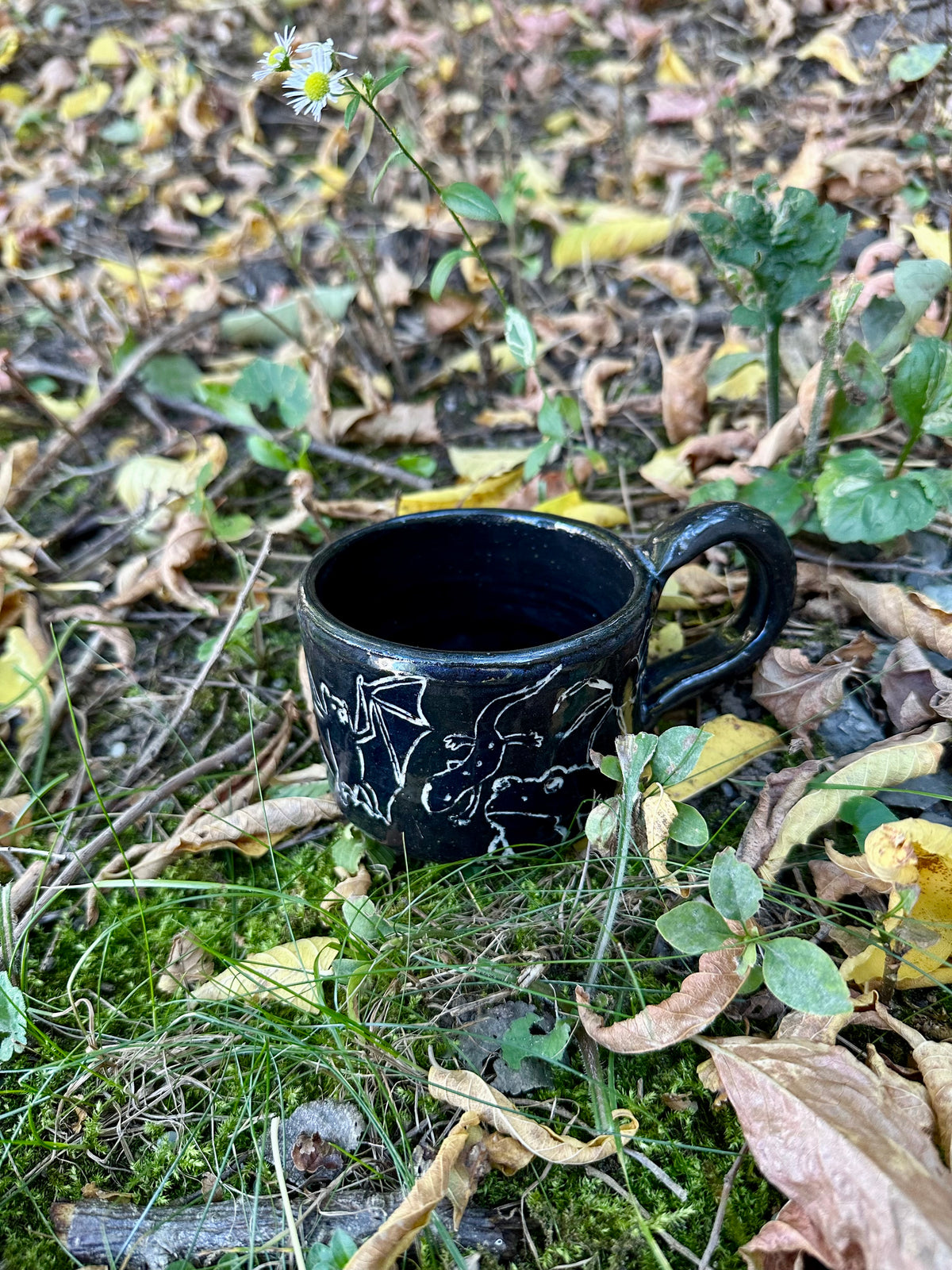 A small black ceramic cup with white line-art illustrations of figures and animals, placed outdoors on a branch surrounded by dry twigs and green leaves.