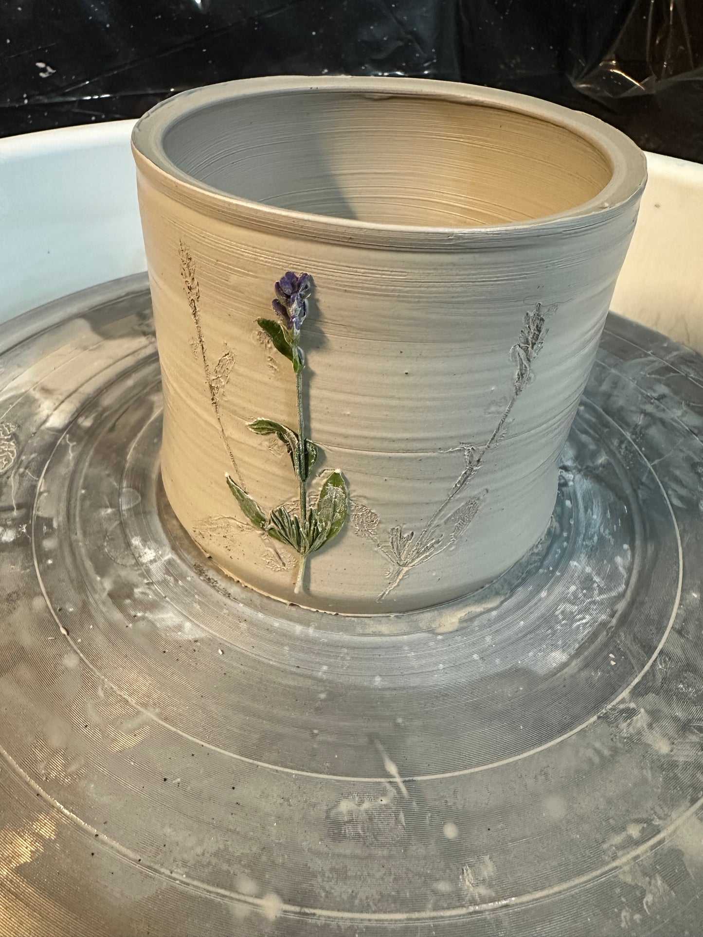 The ceramic cylinder on a pottery wheel when it was still wet; showing the process of real lavender stems being pressed into the clay to make impressions that were later painted.