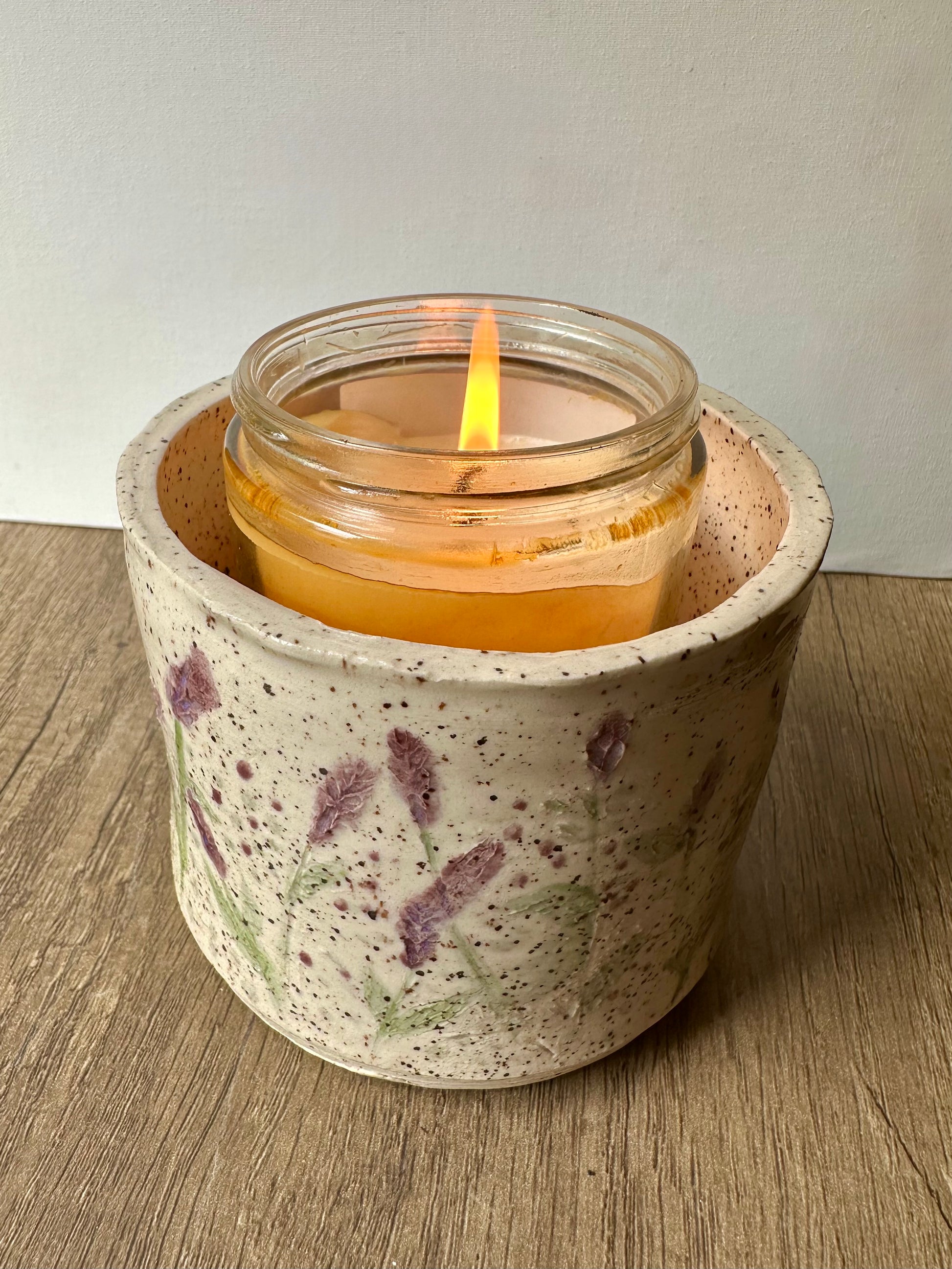 A lit candle inside a speckled white clay cylinder with pressed and painted lavender stems, and purple and green polka dots.