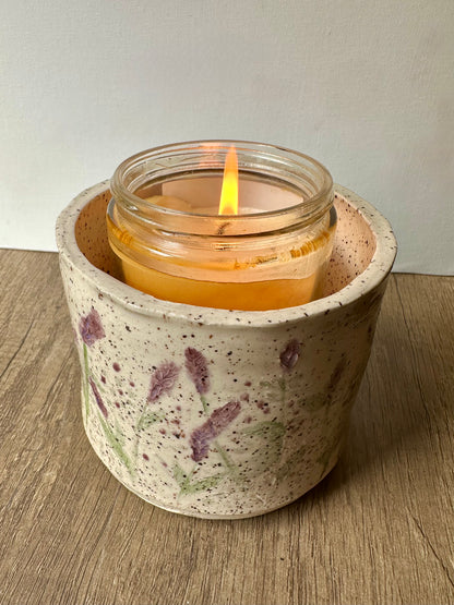 A lit candle inside a speckled white clay cylinder with pressed and painted lavender stems, and purple and green polka dots.