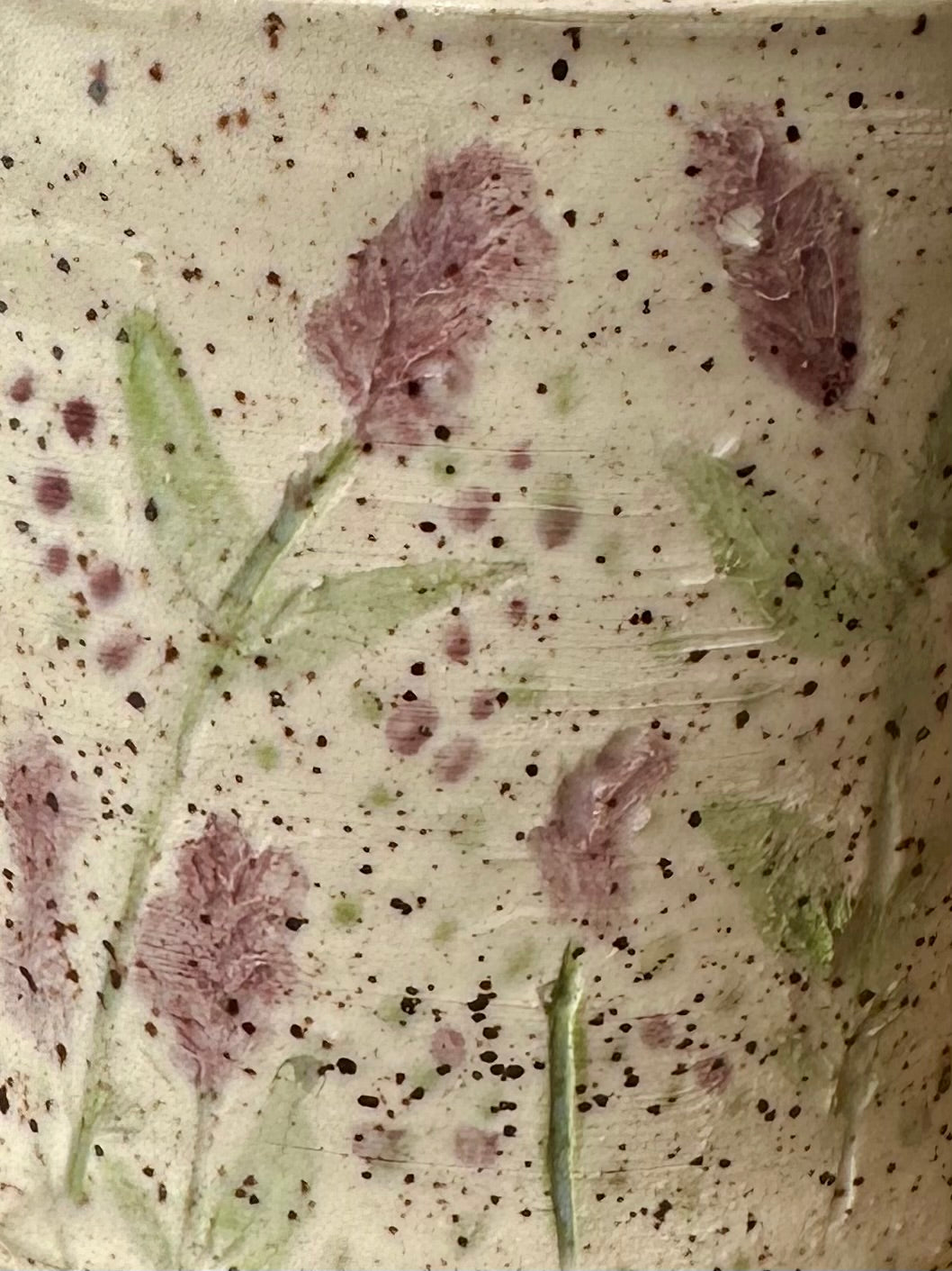 Close-up of a textured white ceramic freckled with small black speckles, with pressed and painted purple lavender stems and leaves.