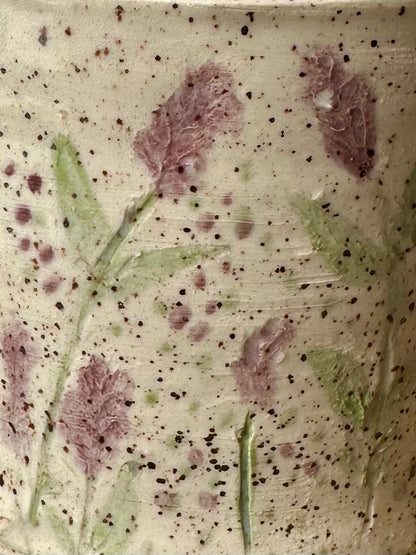 Close-up of a textured white ceramic freckled with small black speckles, with pressed and painted purple lavender stems and leaves.