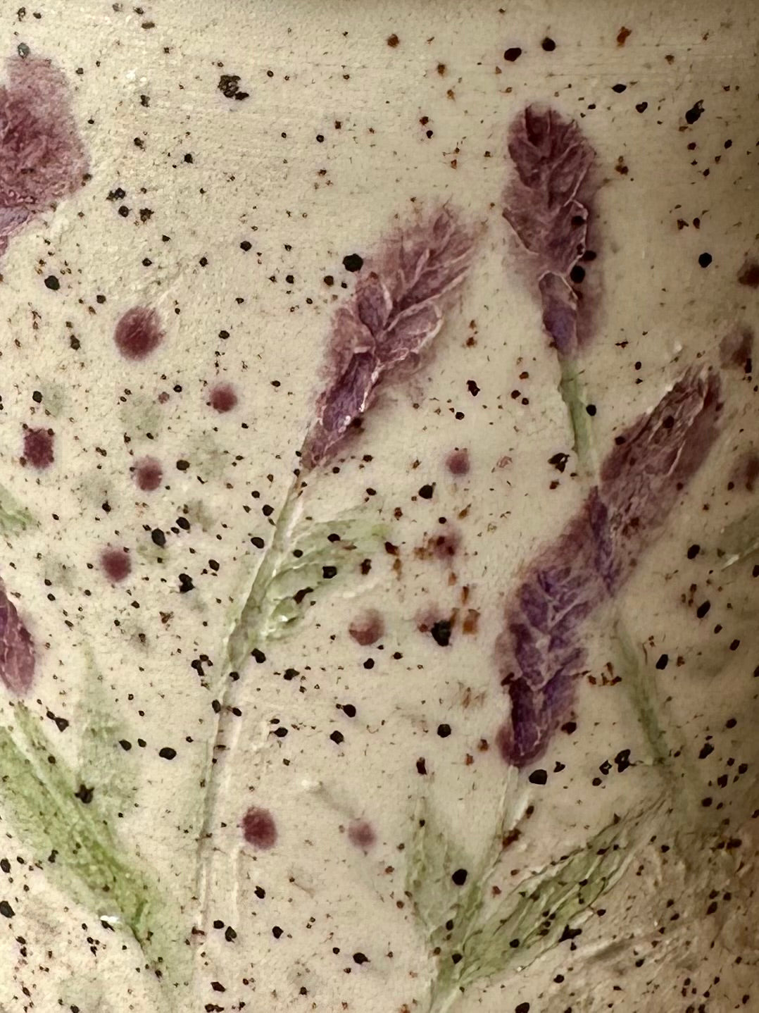 Close-up of a textured white ceramic freckled with small black speckles, with pressed and painted purple lavender stems and leaves.