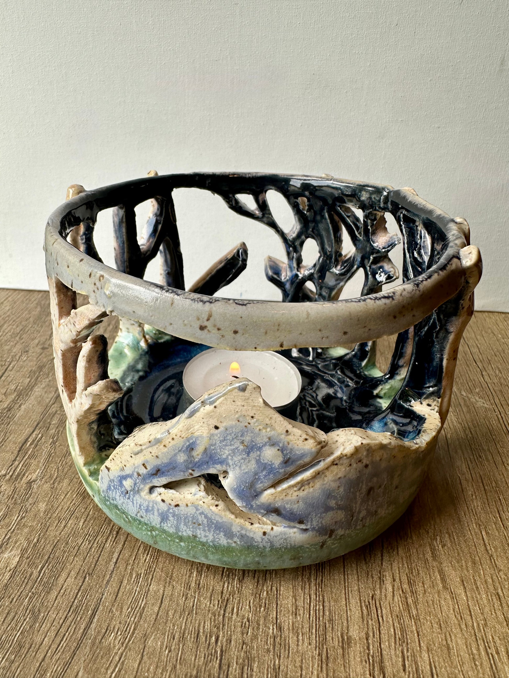Round ceramic candle surround with mountain and birch trees cut out; lit tea light candle placed inside.