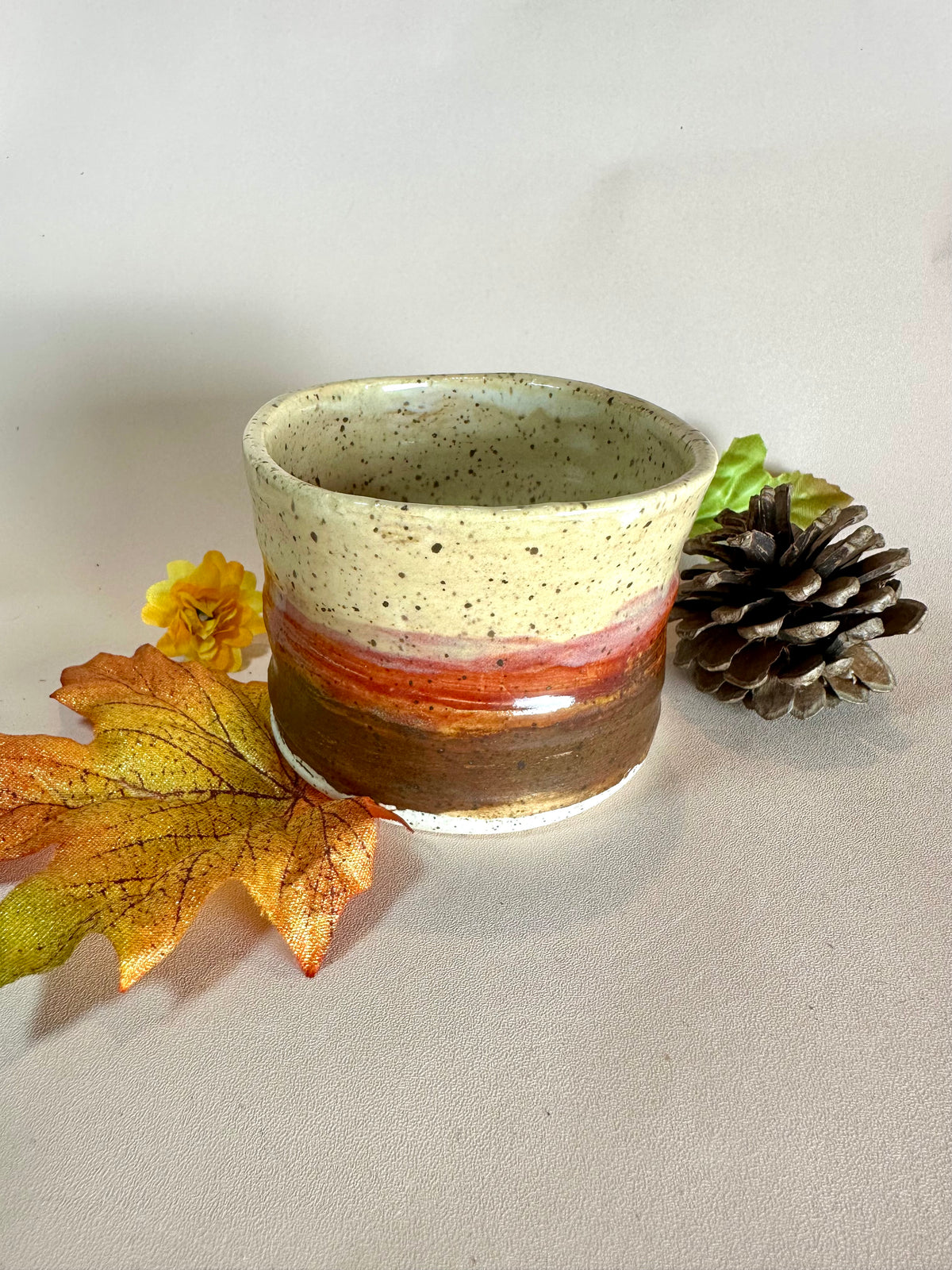 A handmade ceramic cup with a speckled cream top, a red-orange band, and a brown base, styled with autumn leaves, a pinecone, and a small yellow flower on a neutral background.