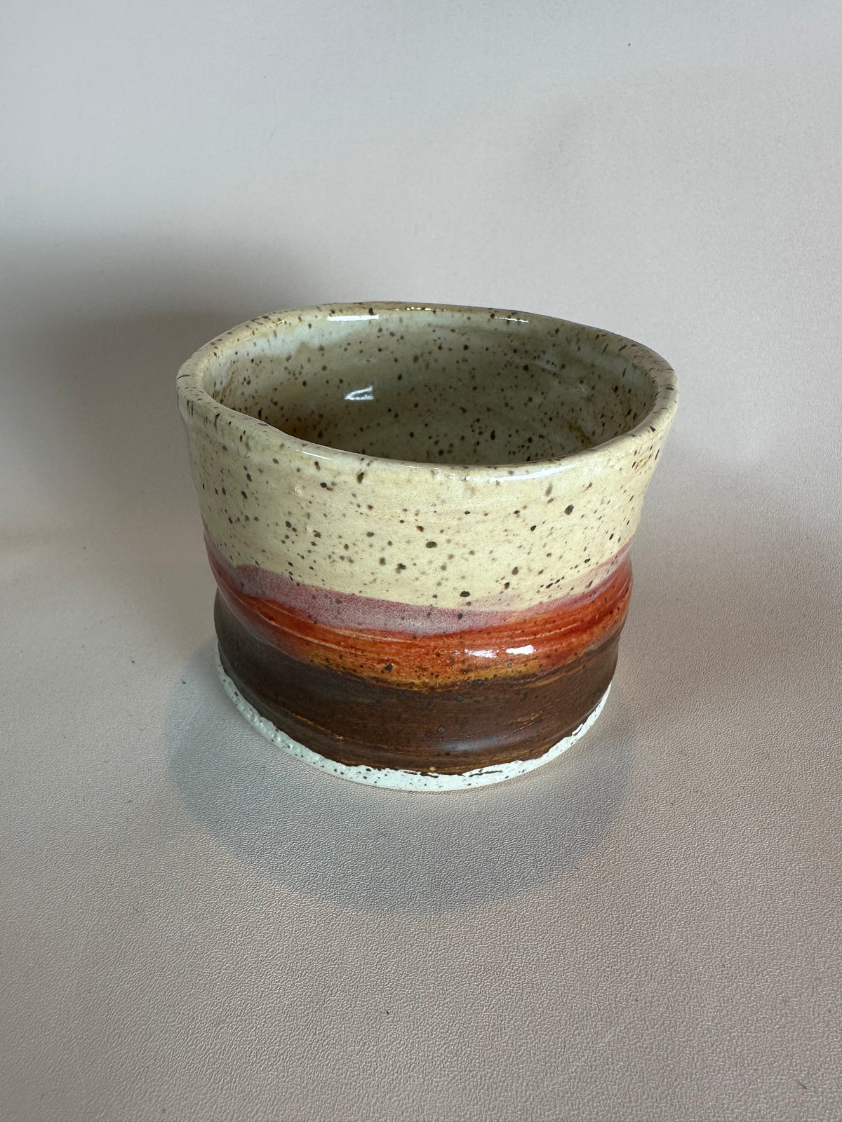 A handmade ceramic cup with a speckled cream top, a red-orange band, and a brown base, styled on a solid neutral background.
