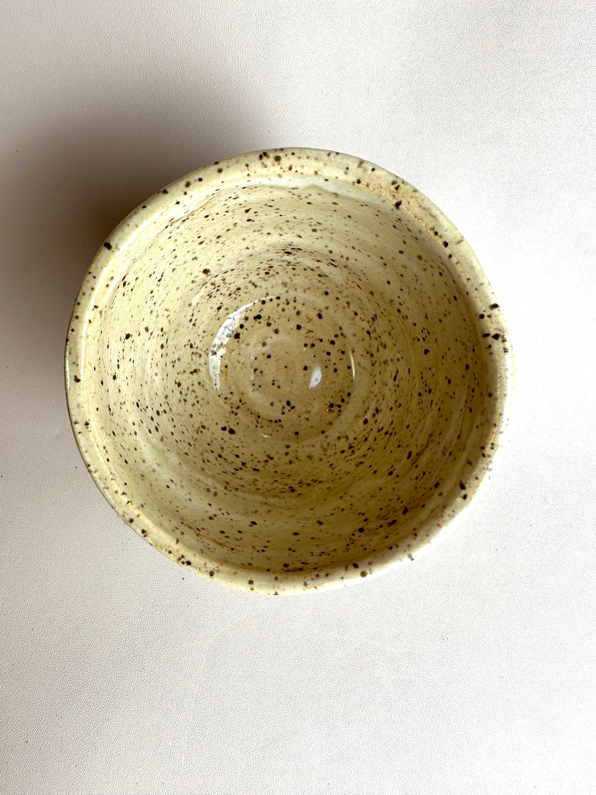 A handmade speckled creamy off-white ceramic cup, photographed from a top-down perspective on a light off-white background.