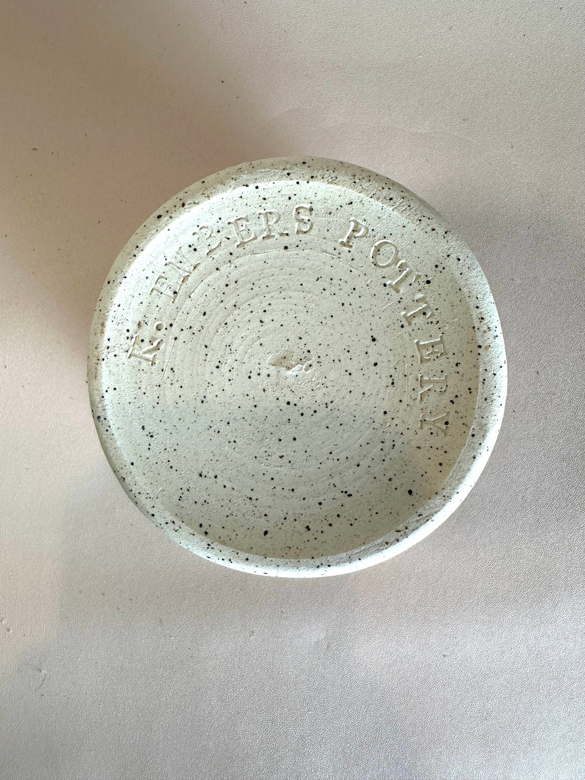 Bottom of a speckled white ceramic cup with "K. Embers Pottery" stamped at the base; set on a solid neutral background.