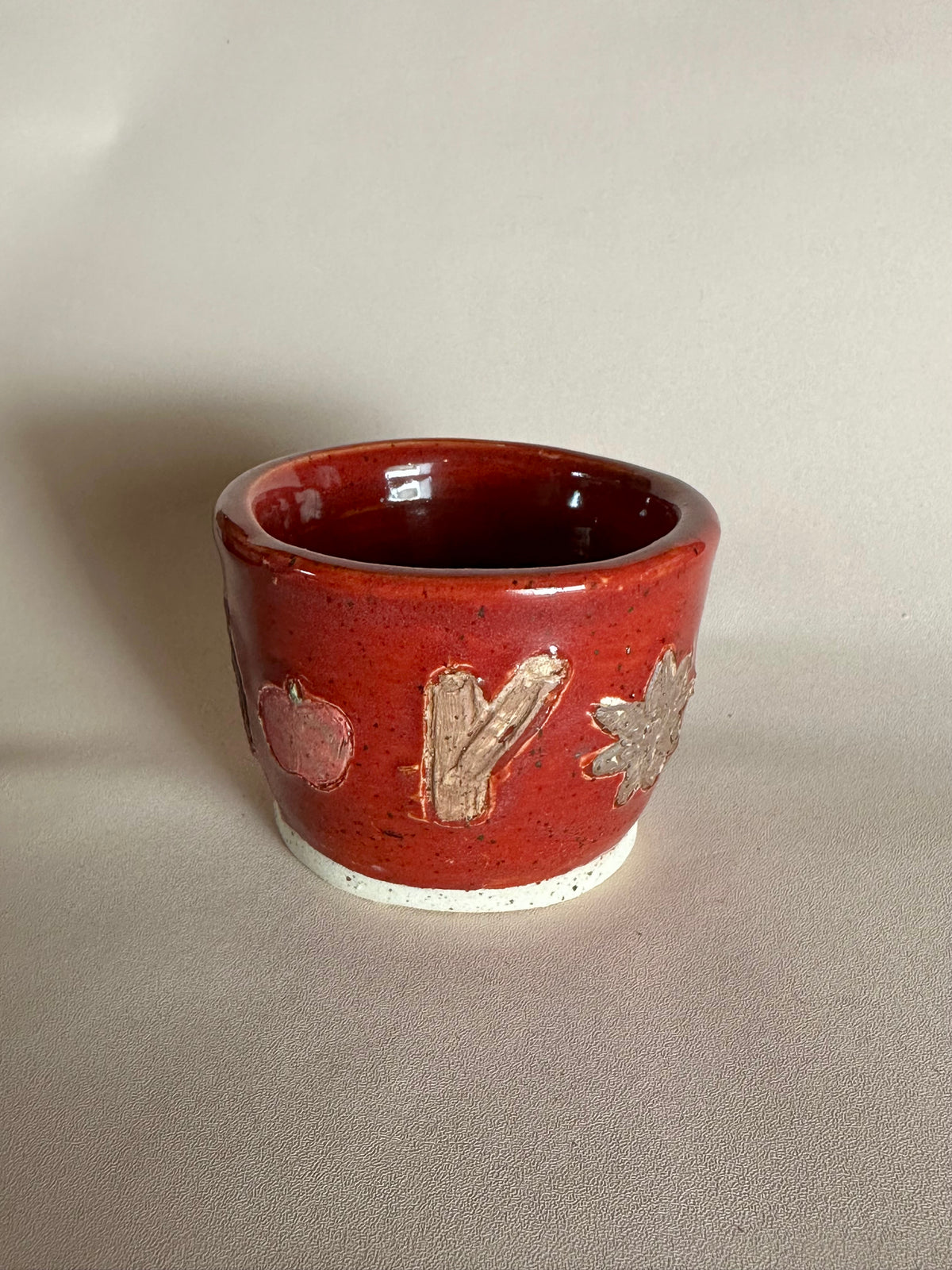 A small handmade ceramic cup with a glossy red glaze and hand-painted apples, cinnamon, and star anise; styled on a neutral background.
