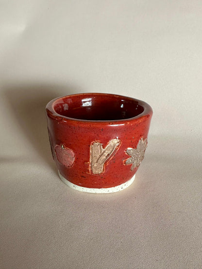 A small handmade ceramic cup with a glossy red glaze and hand-painted apples, cinnamon, and star anise; styled on a neutral background.