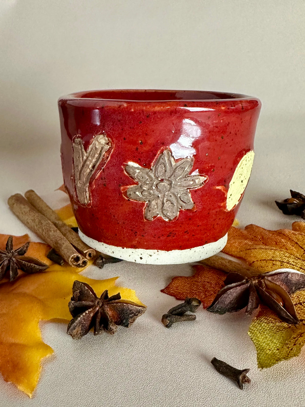 A small handmade ceramic cup with a glossy red glaze and hand-painted apple slice, star anise, and cinnamon; styled with cinnamon sticks, cloves, and fall leaves on a neutral background.
