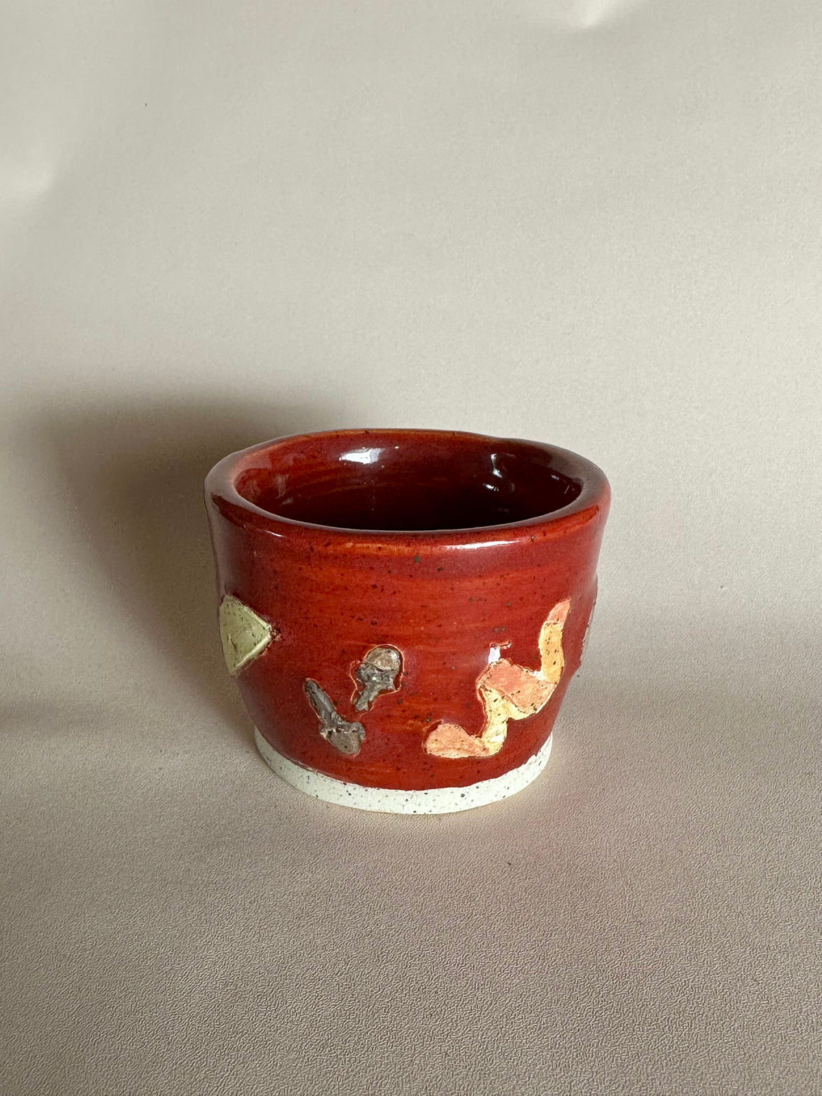 A small handmade ceramic cup with a glossy red glaze and hand-painted apple slice, cloves, and orange peel; styled on a neutral background.