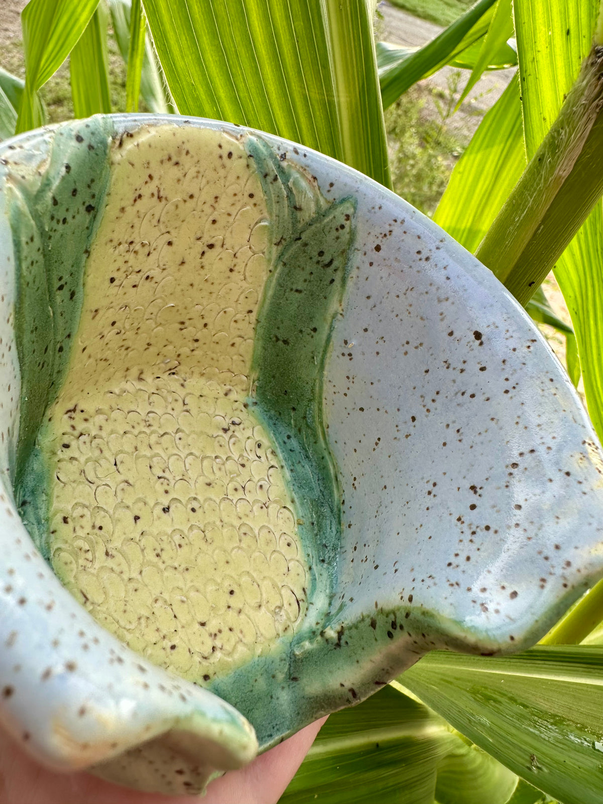 A hand holds a utensil holder themed as a corn stalk against a sky blue base; set in a garden in front of corn stalks.