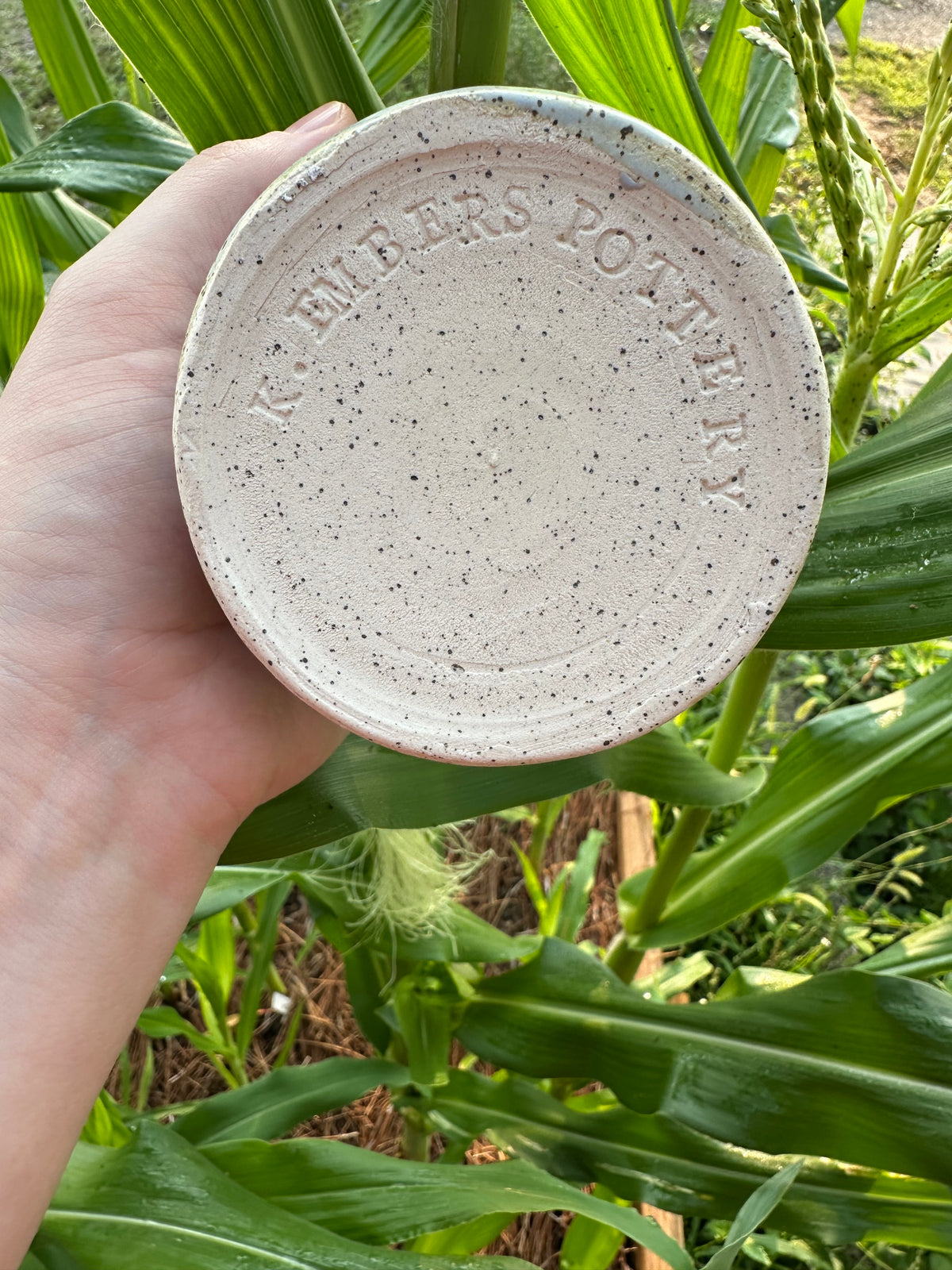 The bottom of a corn-themed utensil holder. The ceramic piece is made of black-speckled white clay, with "K. Embers Pottery" stamped in the base. Set in a garden in front of corn stalks.