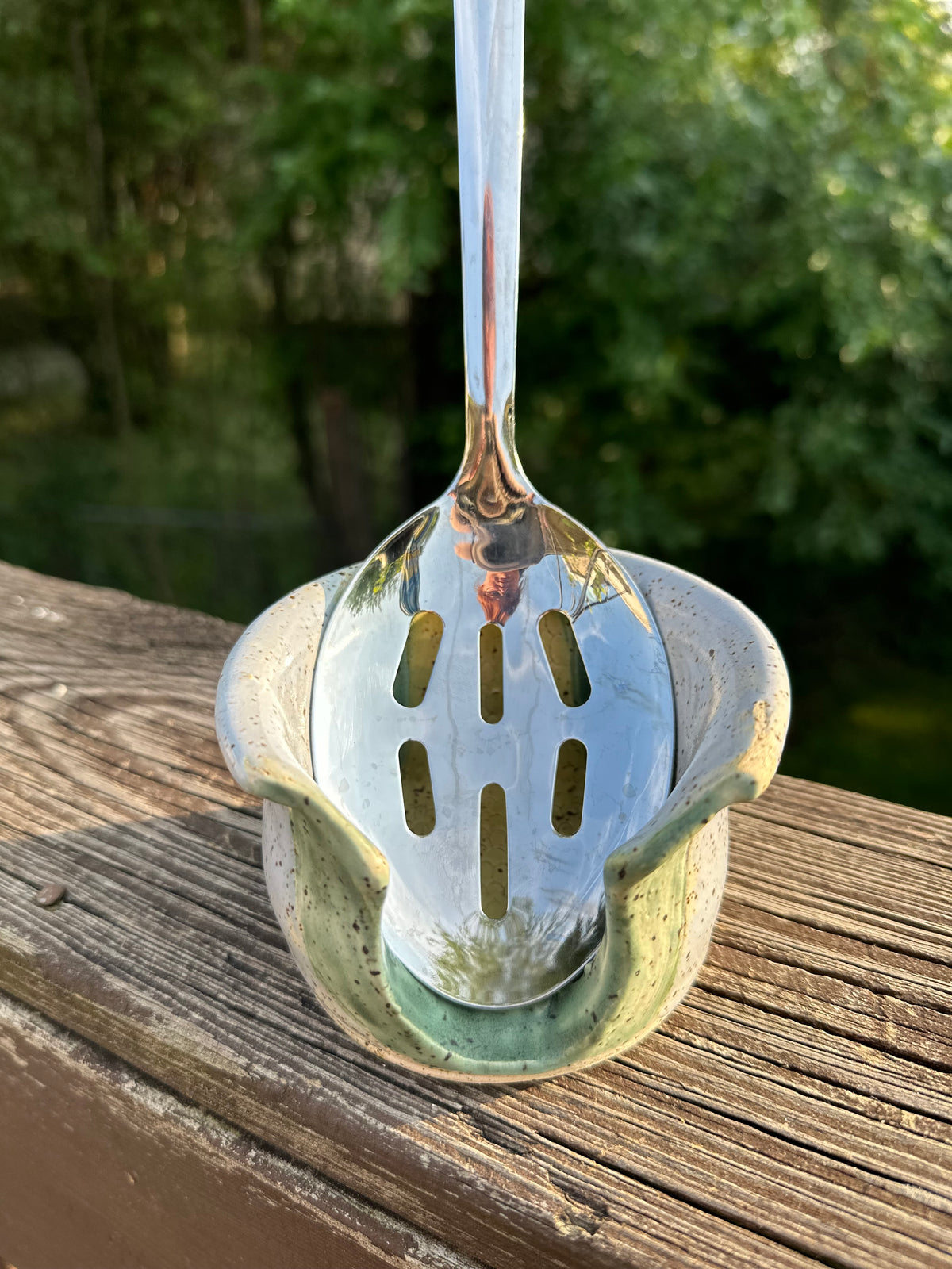 A stainless steel slotted ladle with a long handle, resting inside a small ceramic holder with a green glaze, set on a wooden deck railing with a blurred outdoor background of trees and water.