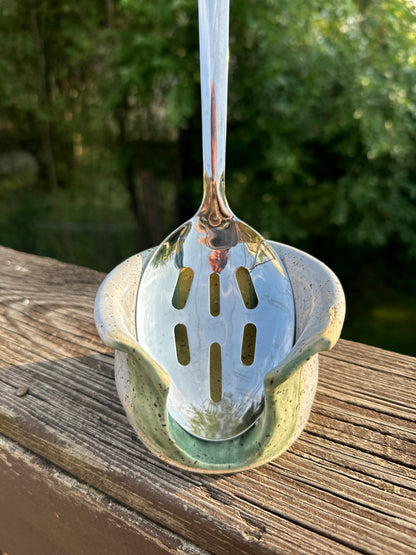 A stainless steel slotted ladle with a long handle, resting inside a small ceramic holder with a green glaze, set on a wooden deck railing with a blurred outdoor background of trees and water.