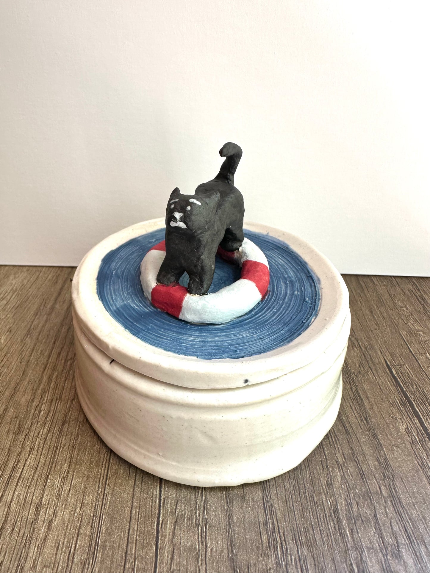 Hero shot of lidded trinket dish. Lid is scared black cat on a life preserver in a pool of water; body is glossy, off-white, lightly speckled.