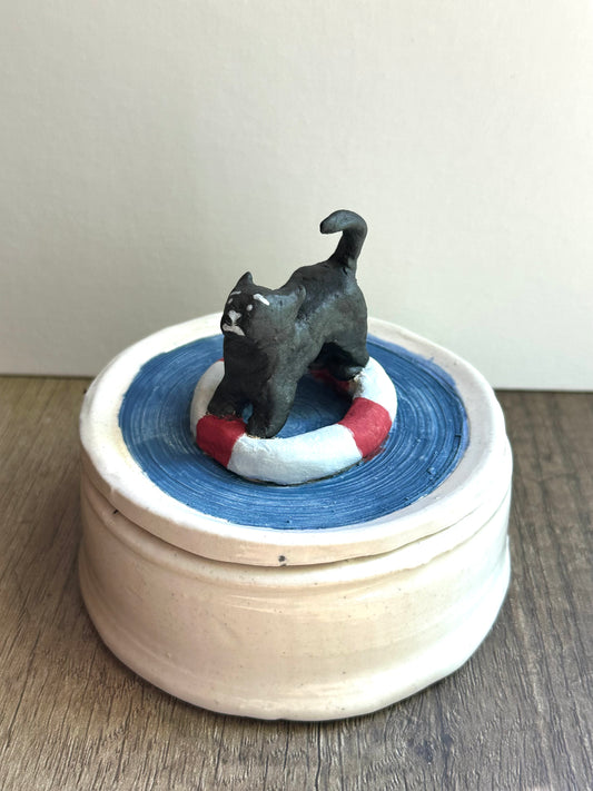 Alternate shot of lidded trinket dish. Lid is scared black cat on a life preserver in a pool of water; body is glossy, off-white, lightly speckled.