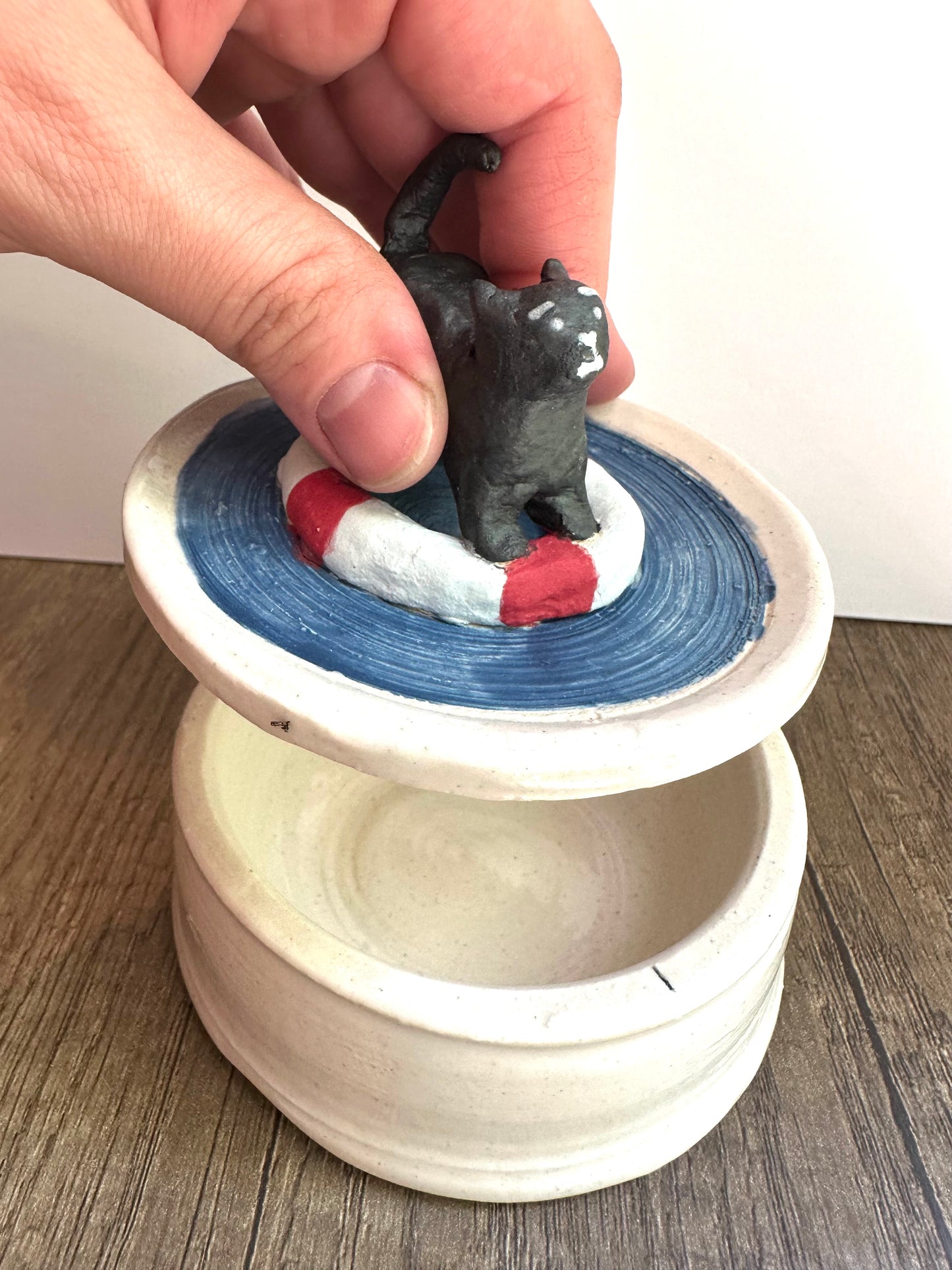 Hand lifting the lid (scared black cat on life preserver in pool of water) of the trinket dish (glossy, off-white, lightly speckled). To show relative in-person scale.