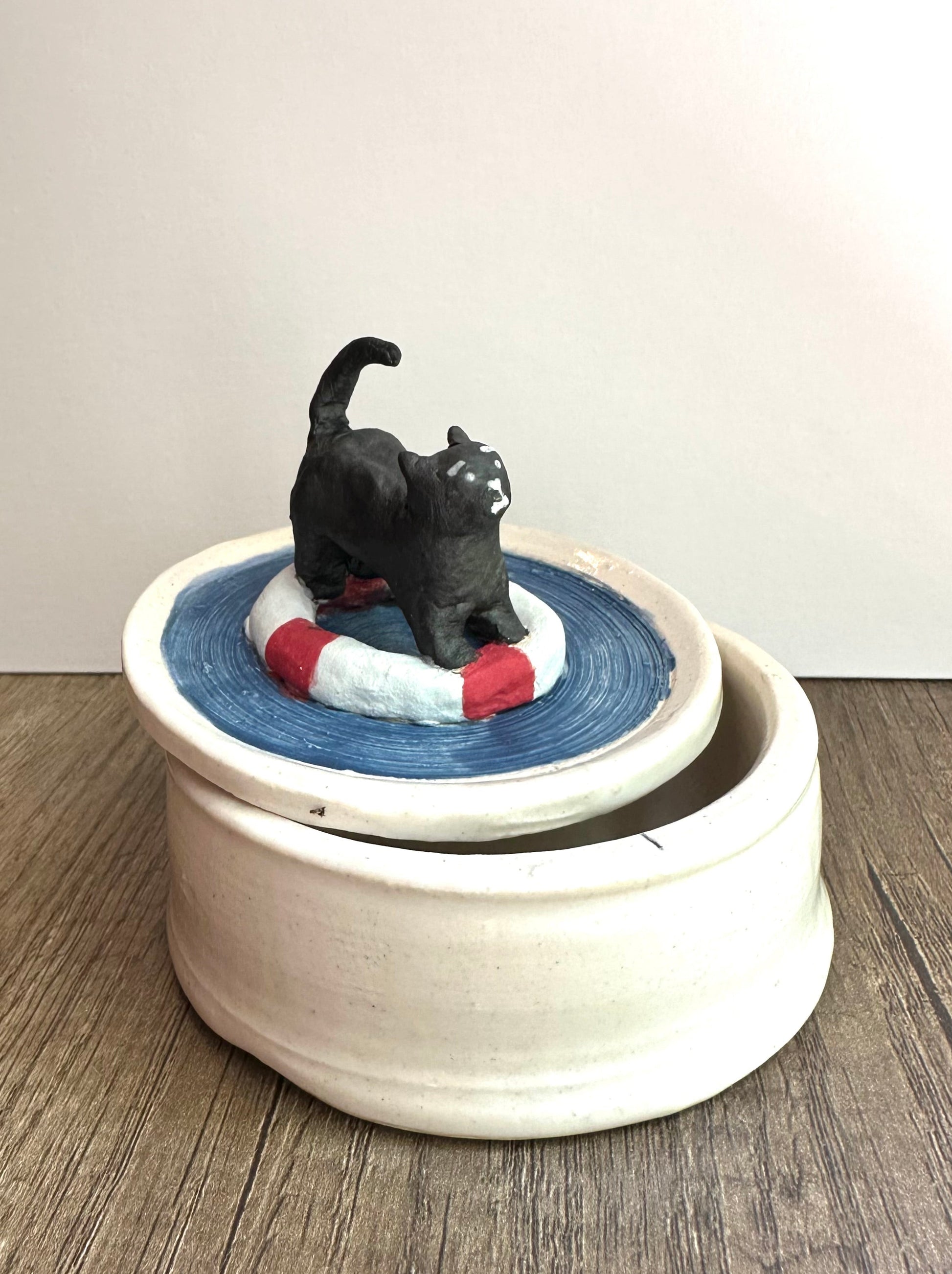 Off-set lid (with black scared cat on life preserver in a pool of water) on top of body of trinket dish (glossy, off-white, lightly speckled).