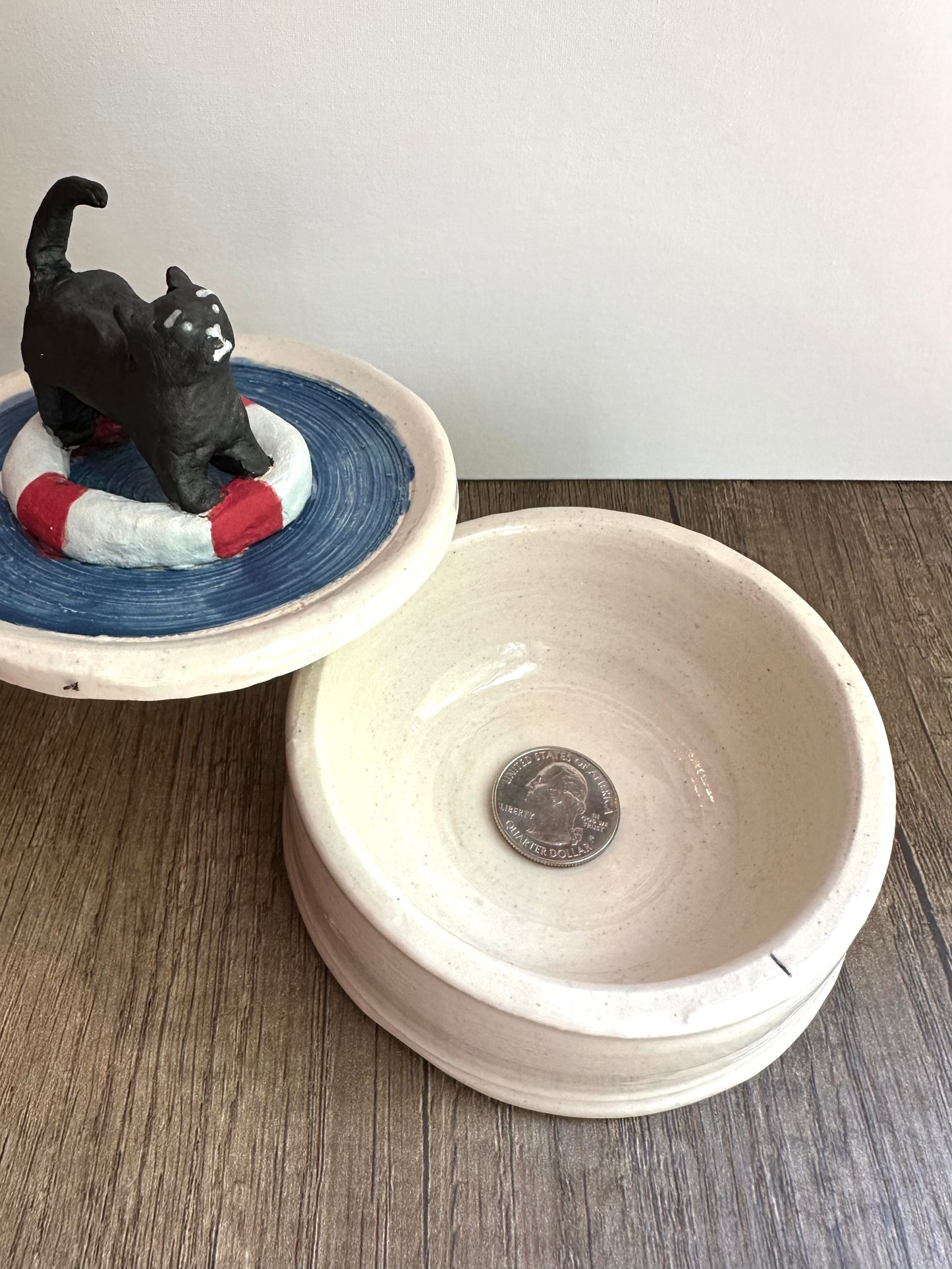 Size reference for the lid and body of the dish. Shows a quarter in the bottom of the body of the dish with the lid (black scared cat on a life preserver on a body of water) off to the side.