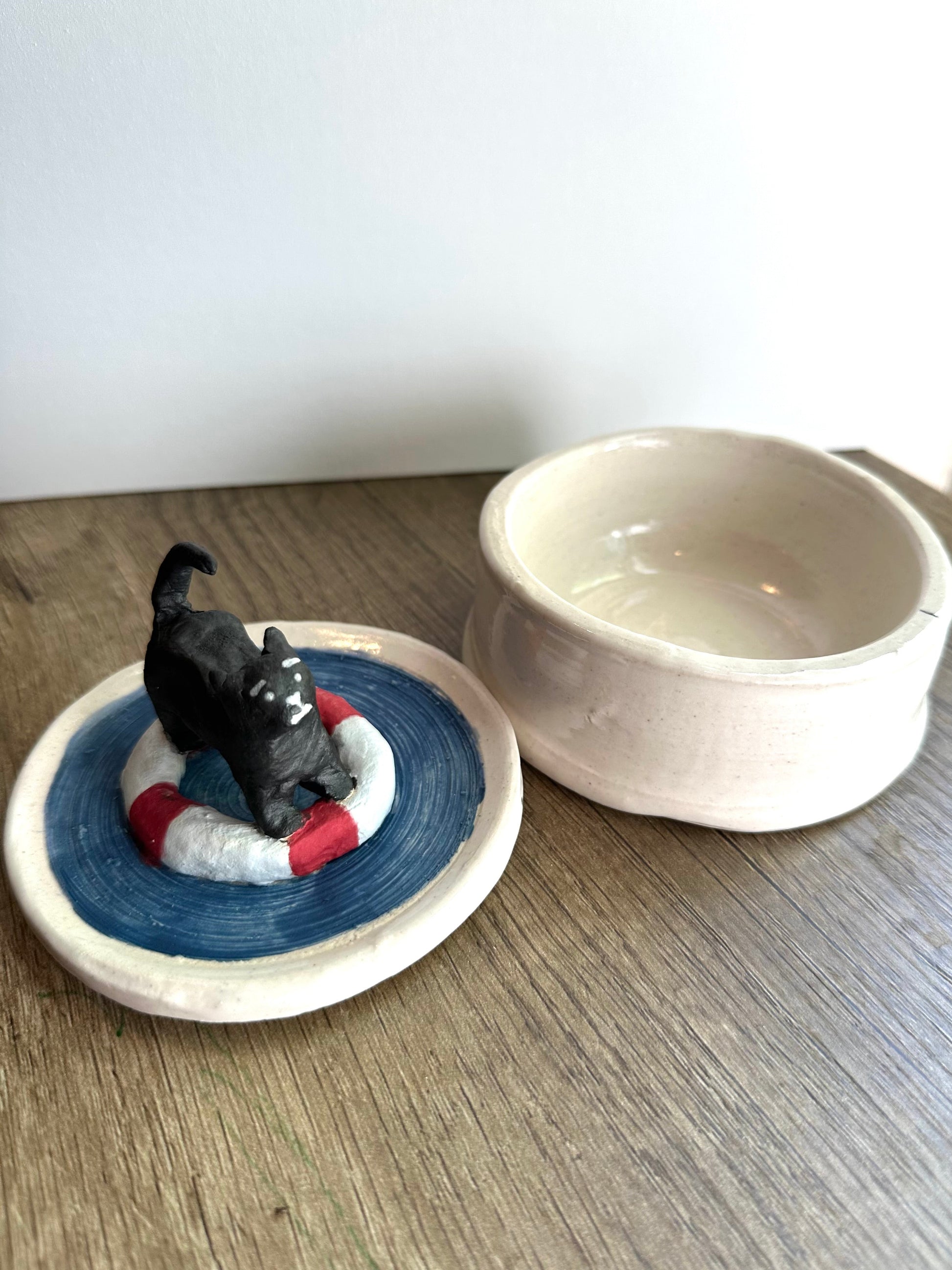 Side by side of the lid (scared black cat on a life preserver in a pool of water) next to the body of the glossy white trinket dish.