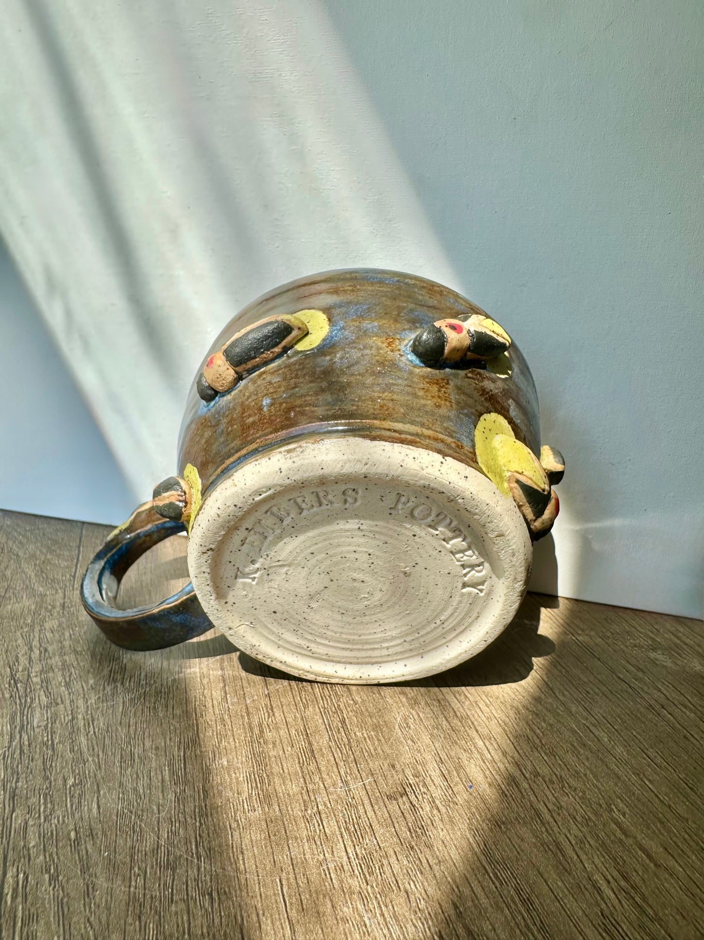 Bronze-colored mug with streaks of inky blue and sculpted fireflies; bottom of mug is bare, showing white clay with black speckles, and "K. Embers Pottery" stamped.