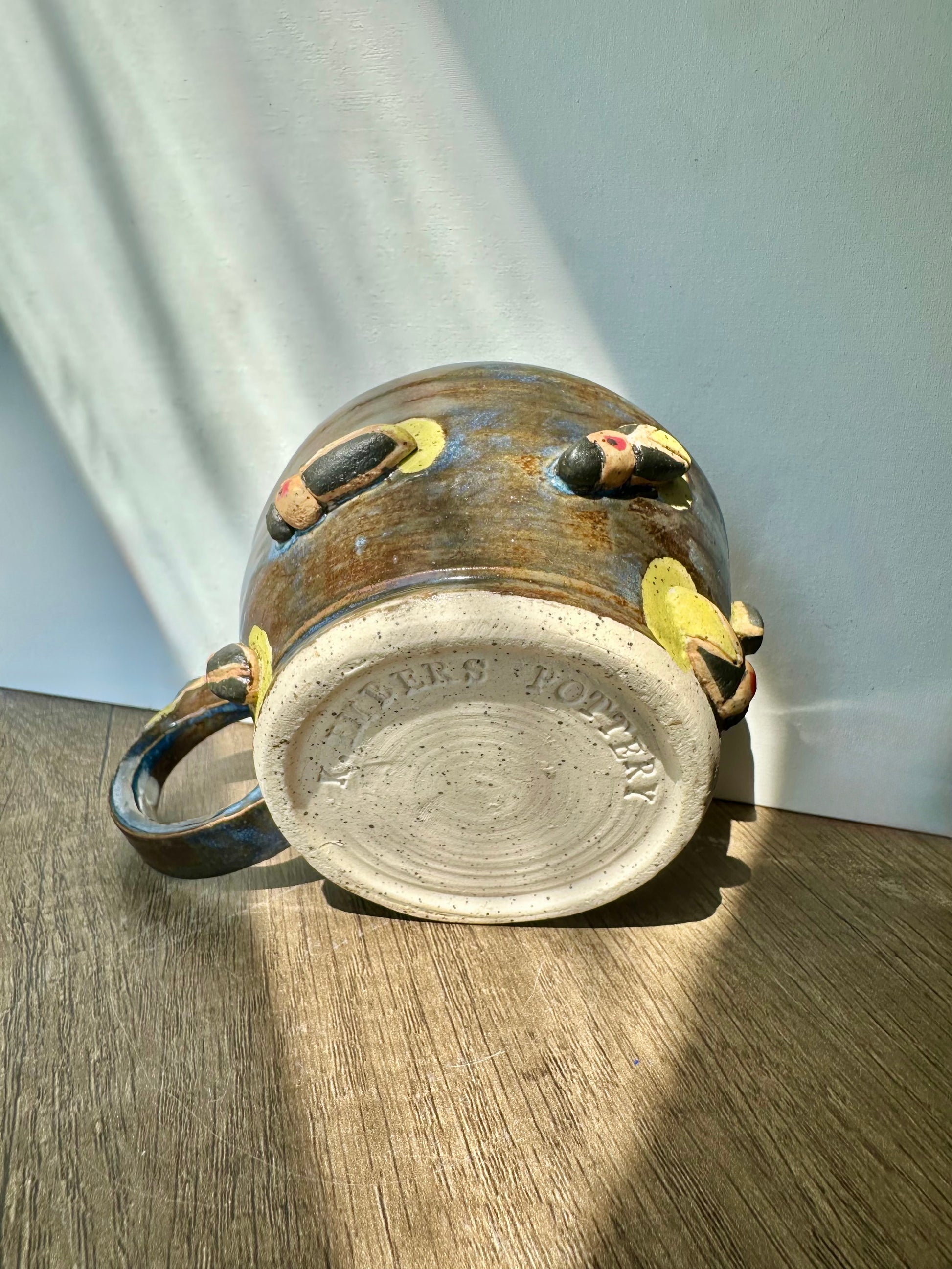 Bronze-colored mug with streaks of inky blue and sculpted fireflies; bottom of mug is bare, showing white clay with black speckles, and "K. Embers Pottery" stamped.
