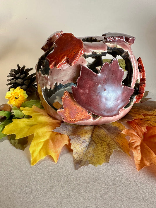 A decorative ceramic sphere with glossy mauve base glaze, featuring maple leaf-shaped cutouts in various shades of red, styled on artificial autumn leaves and a pinecone against a neutral beige background.