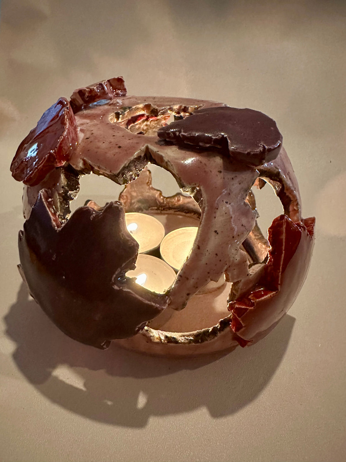 A decorative ceramic sphere with glossy mauve base glaze, featuring maple leaf-shaped cutouts in various shades of red, with three lit tea lights inside, on a neutral beige background.