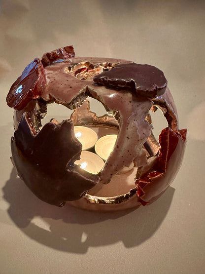 A decorative ceramic sphere with glossy mauve base glaze, featuring maple leaf-shaped cutouts in various shades of red, with three lit tea lights inside, on a neutral beige background.