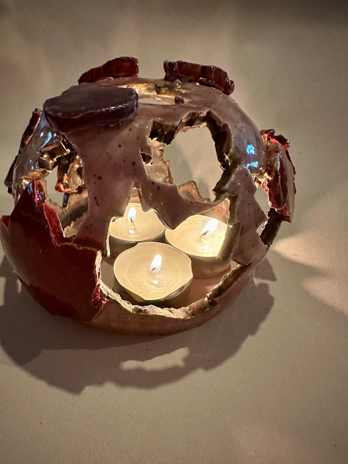 A decorative ceramic sphere with glossy mauve base glaze, featuring maple leaf-shaped cutouts in various shades of red, with three lit tea lights inside, on a neutral beige background.