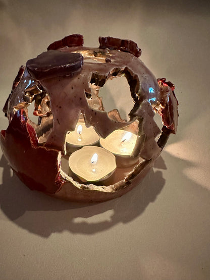 A decorative ceramic sphere with glossy mauve base glaze, featuring maple leaf-shaped cutouts in various shades of red, with three lit tea lights inside, on a neutral beige background.