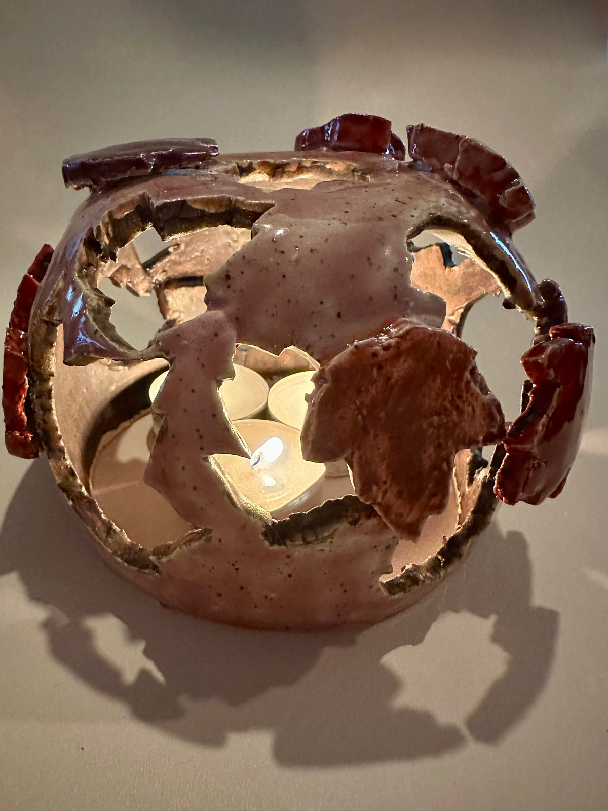 A decorative ceramic sphere with glossy mauve base glaze, featuring maple leaf-shaped cutouts in various shades of red, with three lit tea lights inside, on a neutral beige background.
