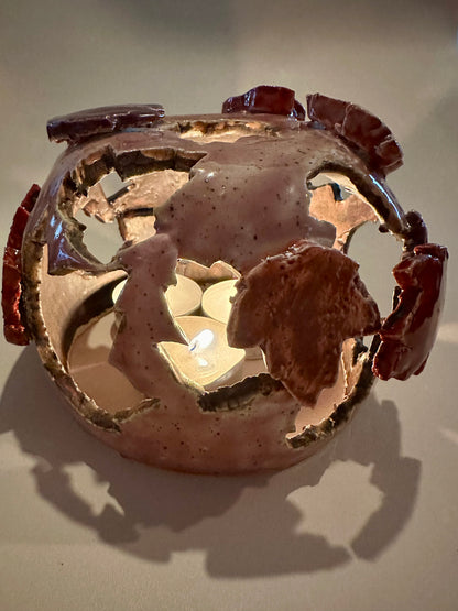 A decorative ceramic sphere with glossy mauve base glaze, featuring maple leaf-shaped cutouts in various shades of red, with three lit tea lights inside, on a neutral beige background.